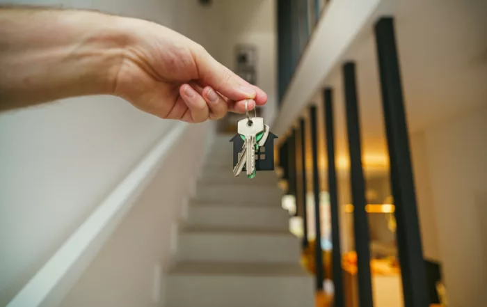 Mano sosteniendo unas llaves con llavero en forma de casa frente a una escalera